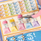 Tweezers placing a flat paint tube sticker inside the miniature art case scene