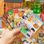Hand holding multiple food journaling sticker sheets with convenience store snacks, drinks, desserts, and Japanese food designs