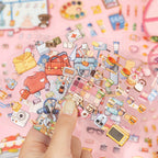 Hand holding transparent sticker sheets with miniature clothing, food, travel, and beauty items