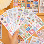Assorted Rainbow and Cloud Stitch sticker sheets held in one hand with colorful embroidered patch-style illustrations