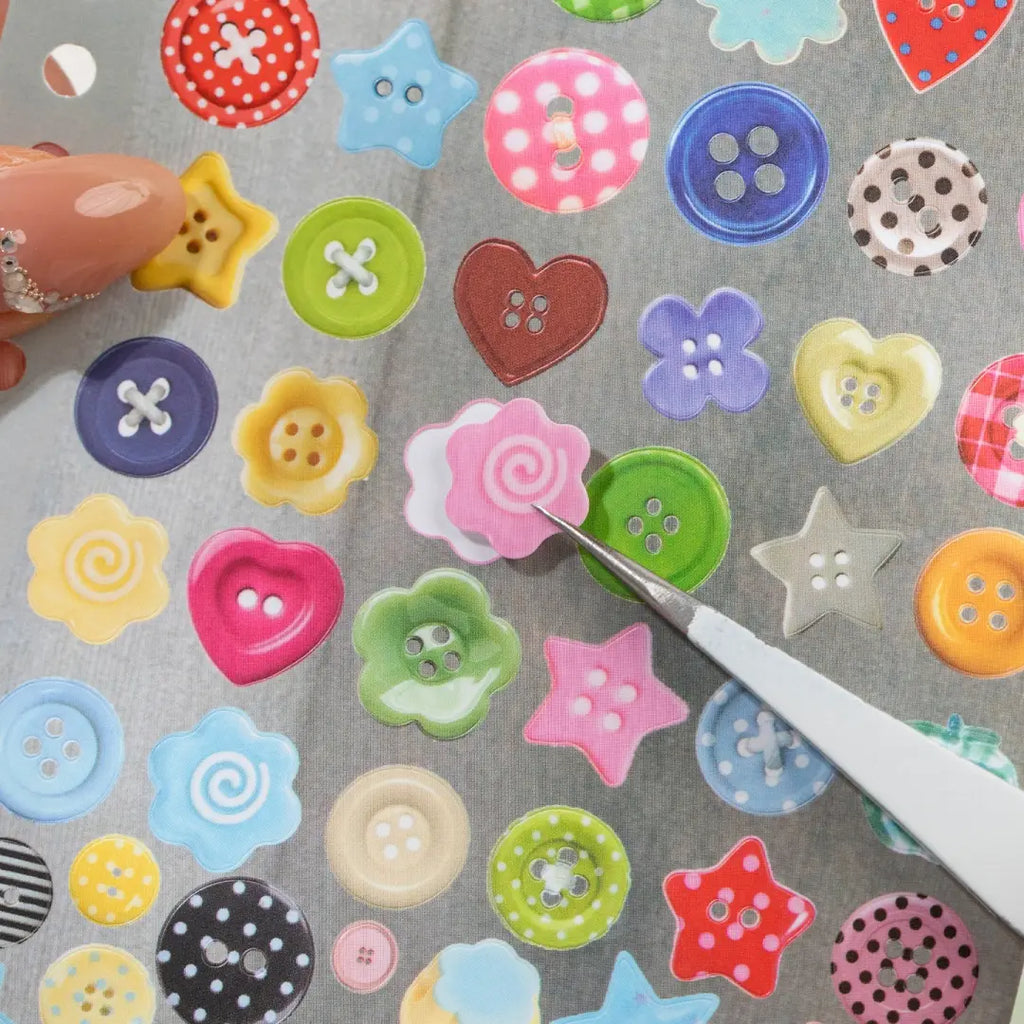 Close-up of colorful button-shaped stickers being lifted from a Strong Valley sticker sheet with tweezers