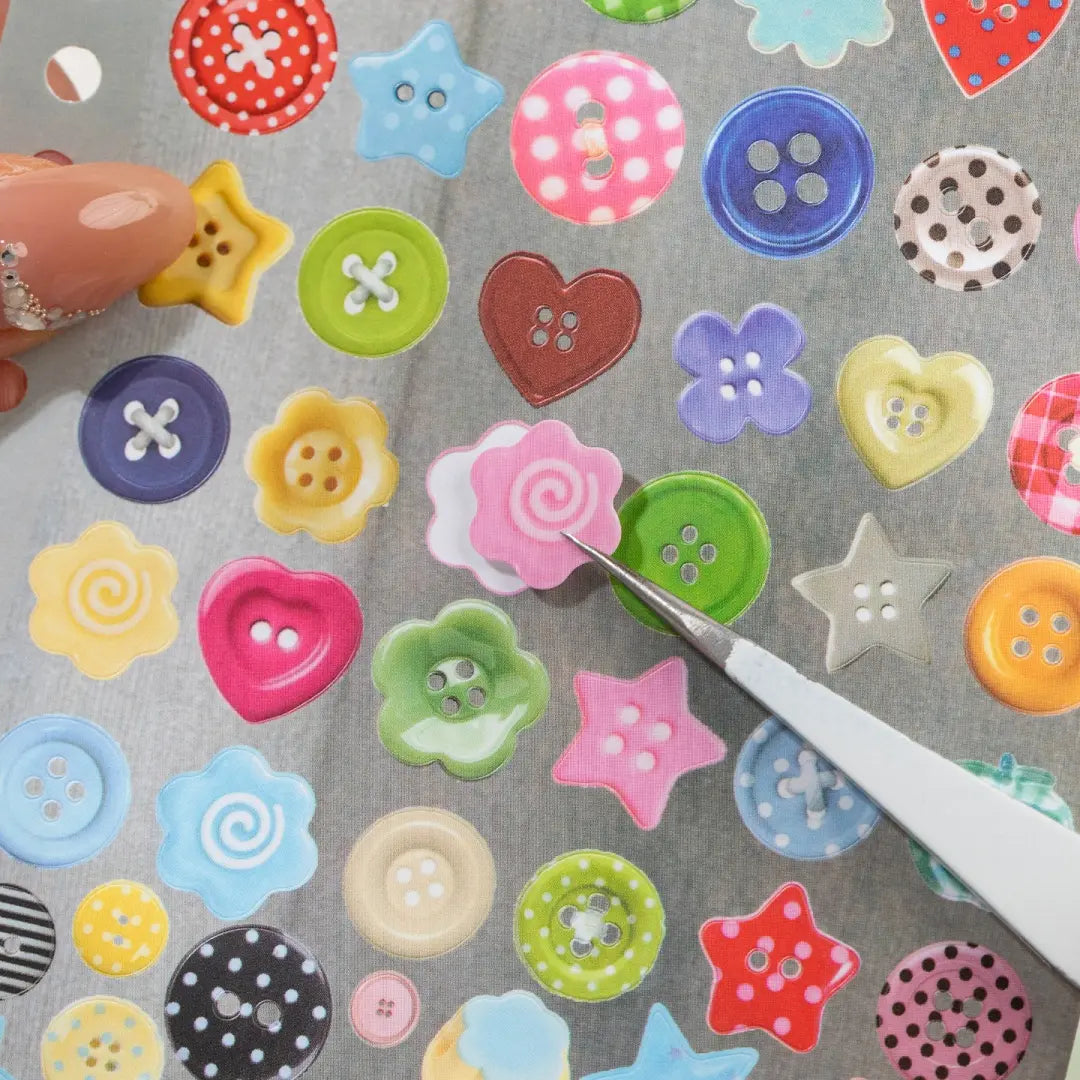 Close-up of colorful button-shaped stickers being lifted from a Strong Valley sticker sheet with tweezers