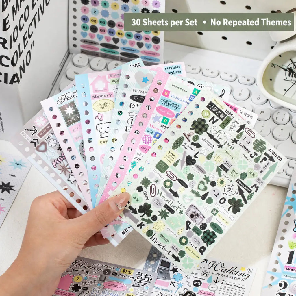 Hand holding several Strong Valley journaling sticker sheets with binder holes, pastel icons, text stickers, labels, arrows, and clover accents